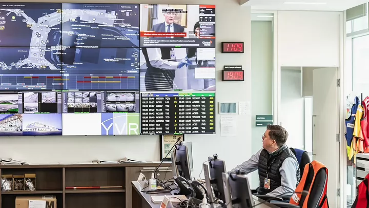 Man sitting at a desk in front of several large digital screens displaying airport information.
