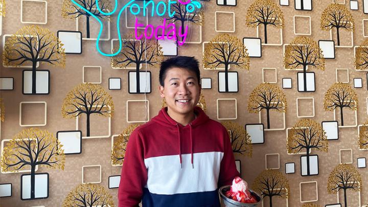 Man is sweat shirt smiling in front of neon sign and holding a shaved ice cream dessert.