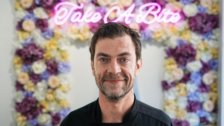 Man smiling in front of flowery neon sign that says Take A Bite.