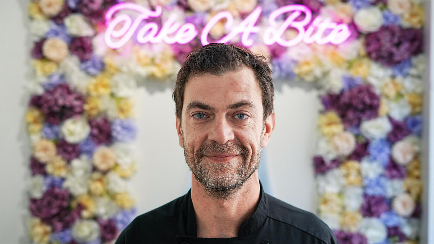 Man smiling in front of flowery neon sign that says Take A Bite.