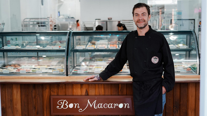 Man wearing black apron standing in front of glass display case filled with plates of macarons and a sign below the display case that reads Bon Macaron