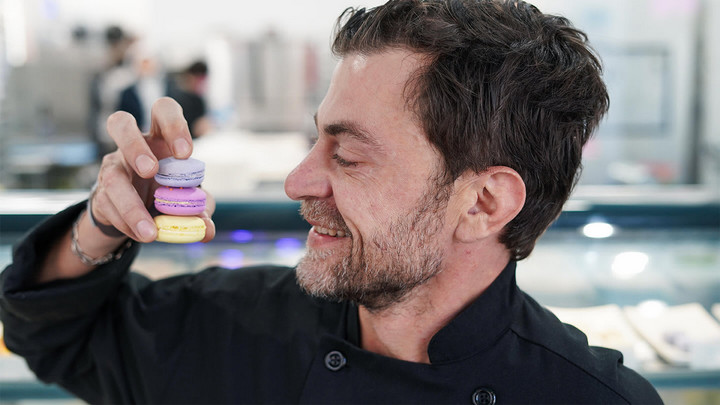 Man smiling as he looks at three macarons he's holding that are stacked on top of one another.