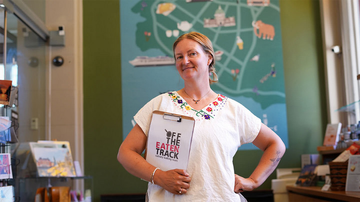 Woman standing in a shop holding a clipboard that says Off the Eaten Track Culinary Walking Tours