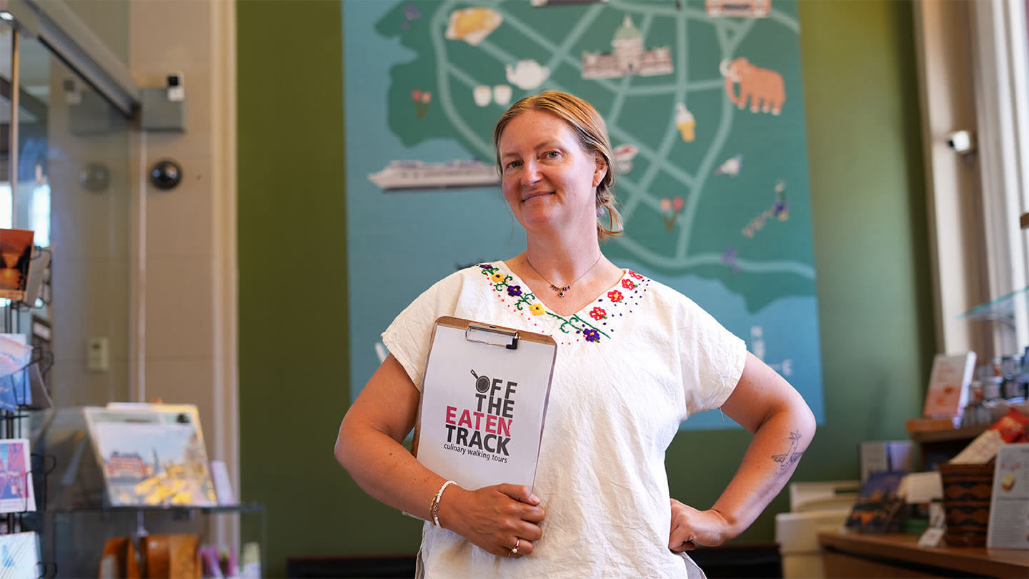 Woman standing in a shop holding a clipboard that says Off the Eaten Track Culinary Walking Tours