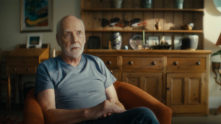 Older man with white facial hair sitting in a chair with a wooden desk and credenza behind him. 