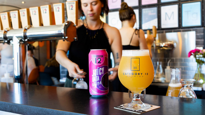 A beer can of Juicy Data IPA beside a full beer glass, with a server and beer taps in the background.