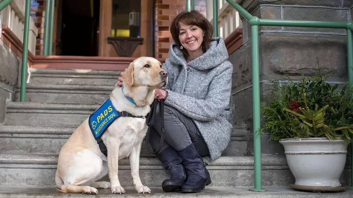 A woman on a set of stairs smiling with an assistance dog.
