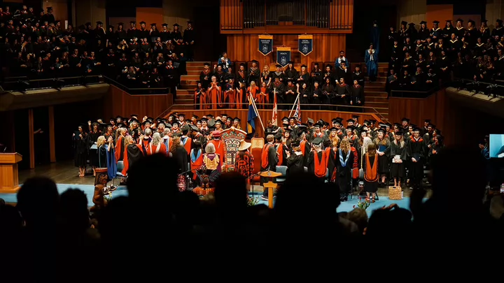 Silhouette of audience standing and applauding as university graduates leave the stage of their graduation ceremony. 