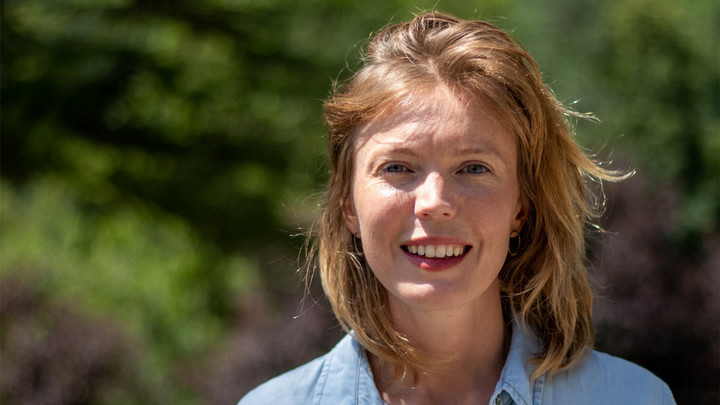 Woman in denim shirt smiling in front of foliage.