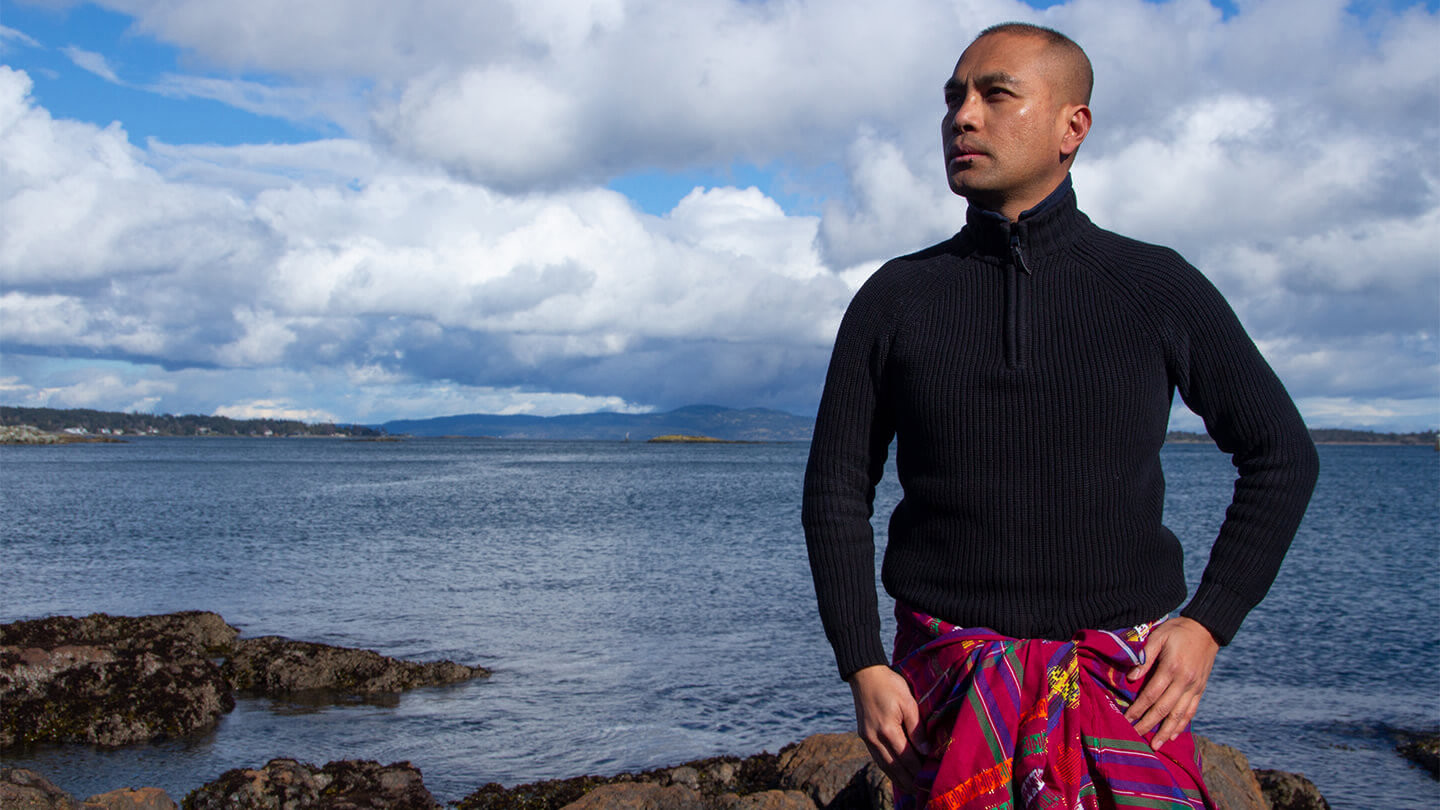 Man with shaved head and colourful pants standing in front of the ocean.