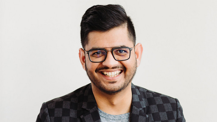 Dark haired man with thin facial hair in dark-framed glasses smiling and wearing a checkered jacket with grey t-shirt.