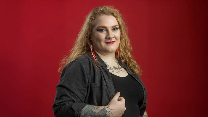 Person with long blond hair, septum piercing, beaded earrings and tattoos on arms and chest.