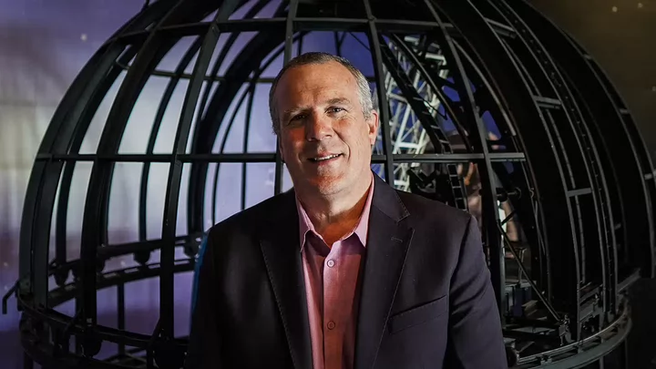 A man in a jacket and dress shirt standing in front of a metal sphere.
