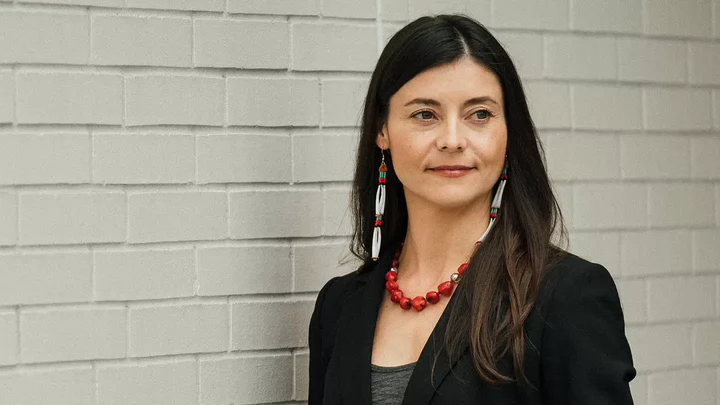 A woman with long hair and beaded earrings standing in front of a pale brick wall.