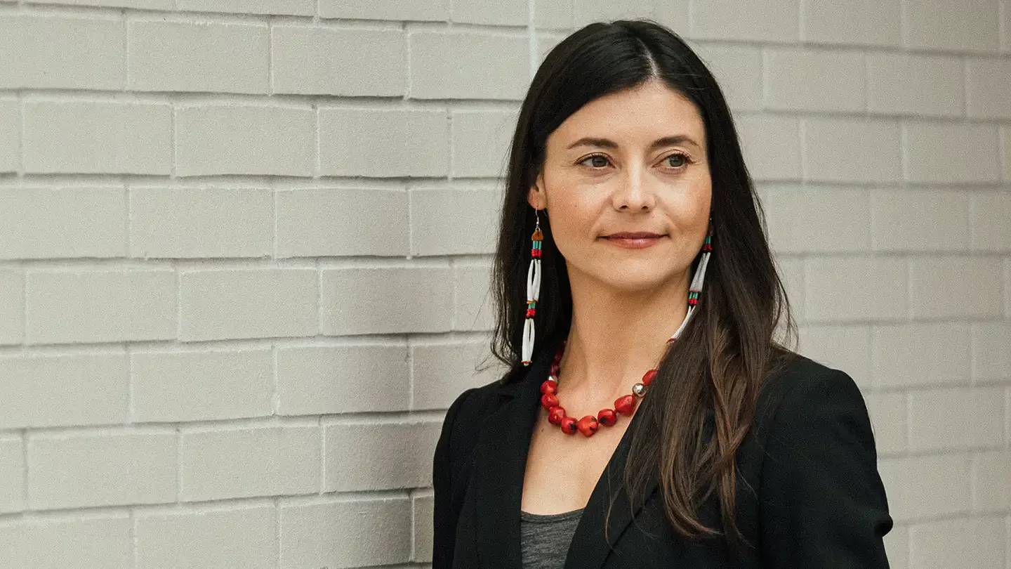 A woman with long hair and beaded earrings standing in front of a pale brick wall.