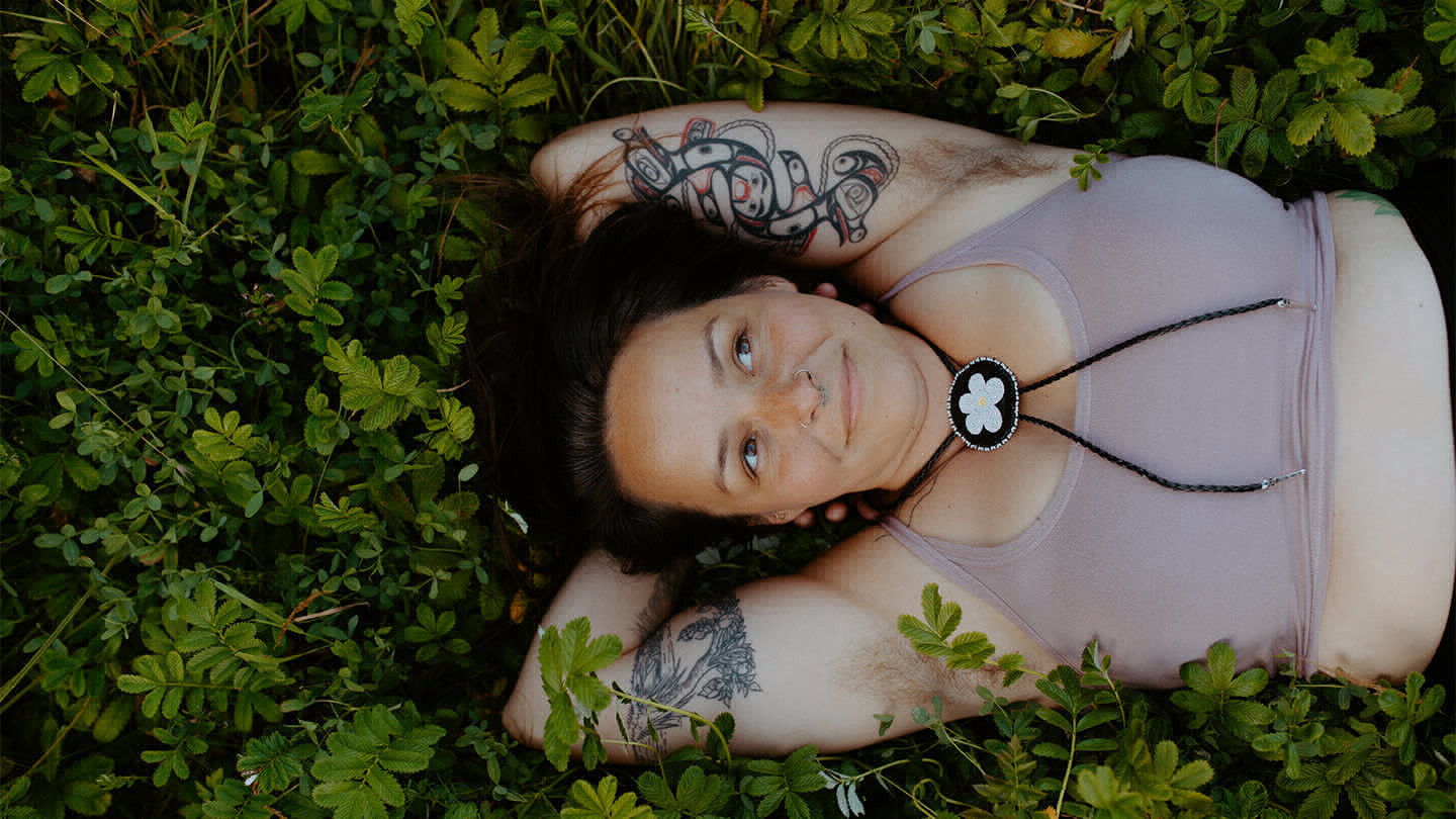 Person with Indigenous tattoos on their arms and beaded necklace lying in a meadow with hands behind their head. 
