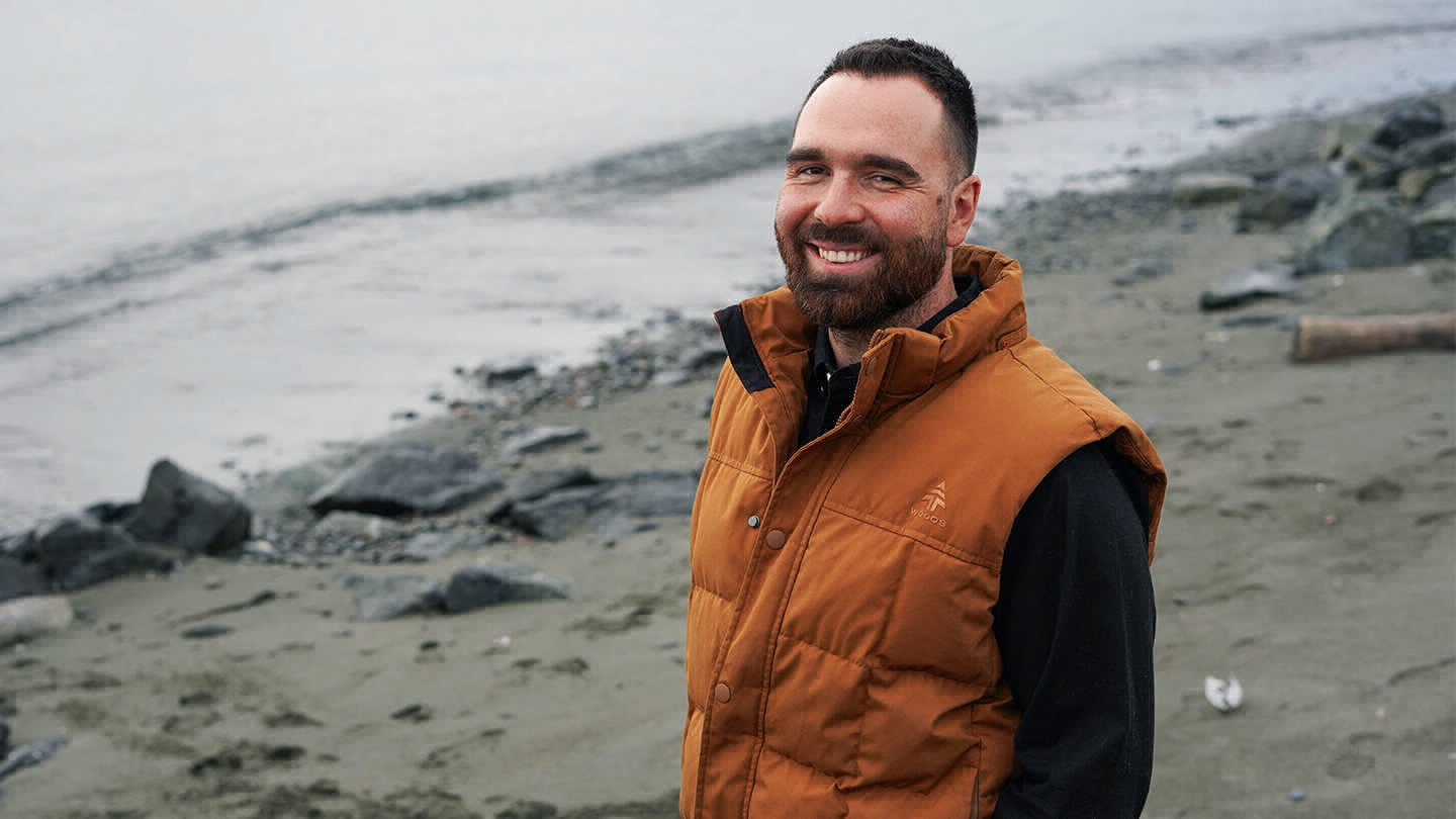 Man with facial hair and wearing puffy vest smiling and standing on a beach.