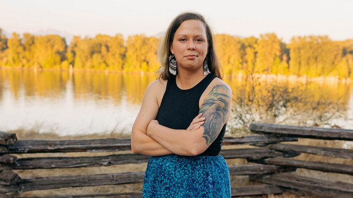 Person wearing beaded earrings standing arms crossed in front of a lake or river.
