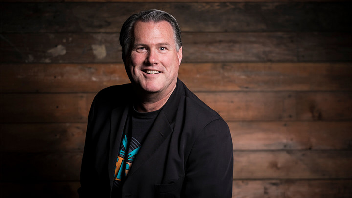 Man smiling while wearing black jacket and T-shirt in front of wood-slatted backdrop.