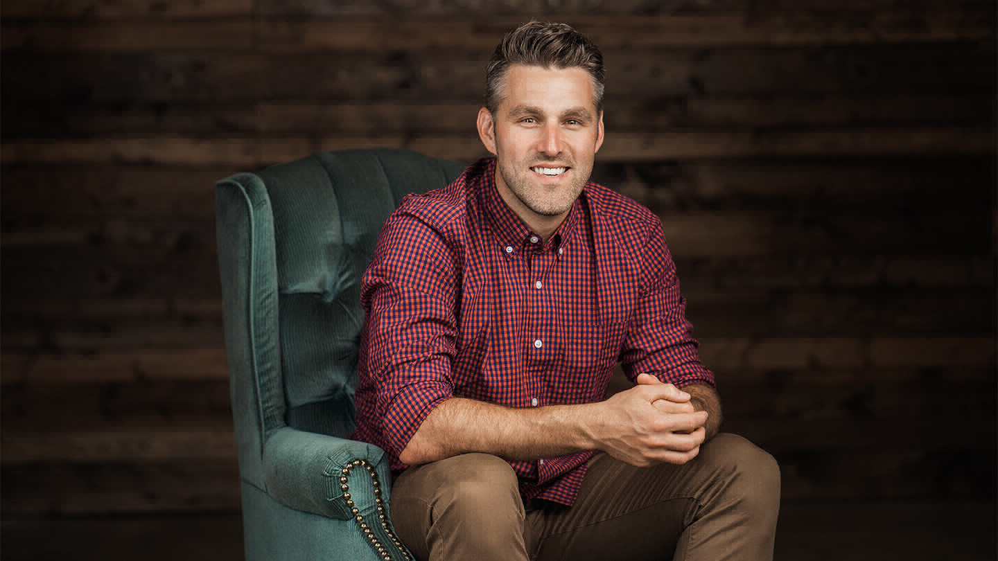 Man with facial stubble wearing a checkered button-up shirt and sitting on a couch.