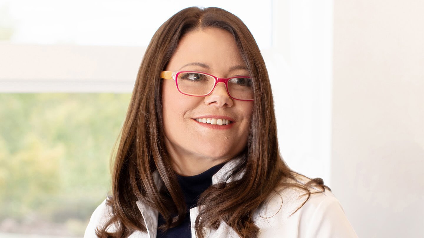 Person with shoulder length hair, wearing glasses and a lab coat smiling.