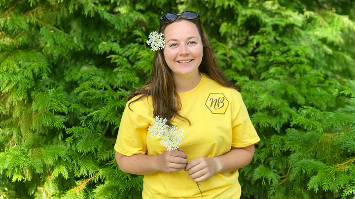 Woman smiling with flowers in her hair while standing in front of green trees and shrubbery.