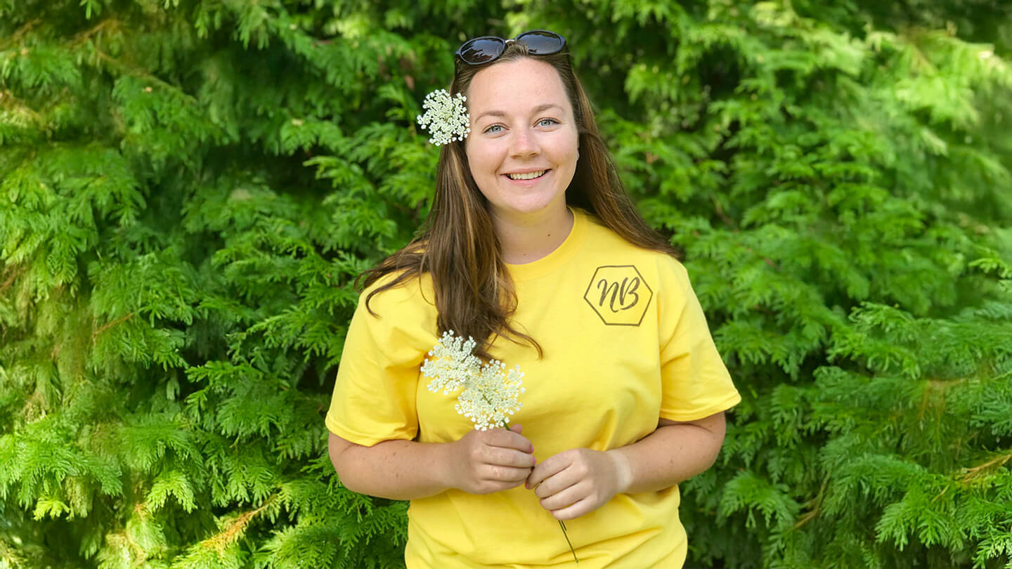Woman smiling with flowers in her hair while standing in front of green trees and shrubbery.