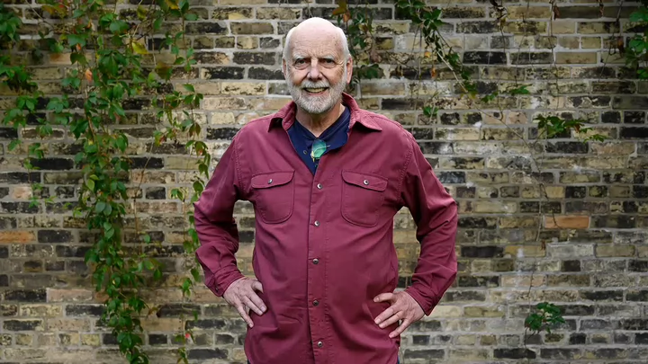 A man with white beard smiling and standing in front of vine covered brick wall