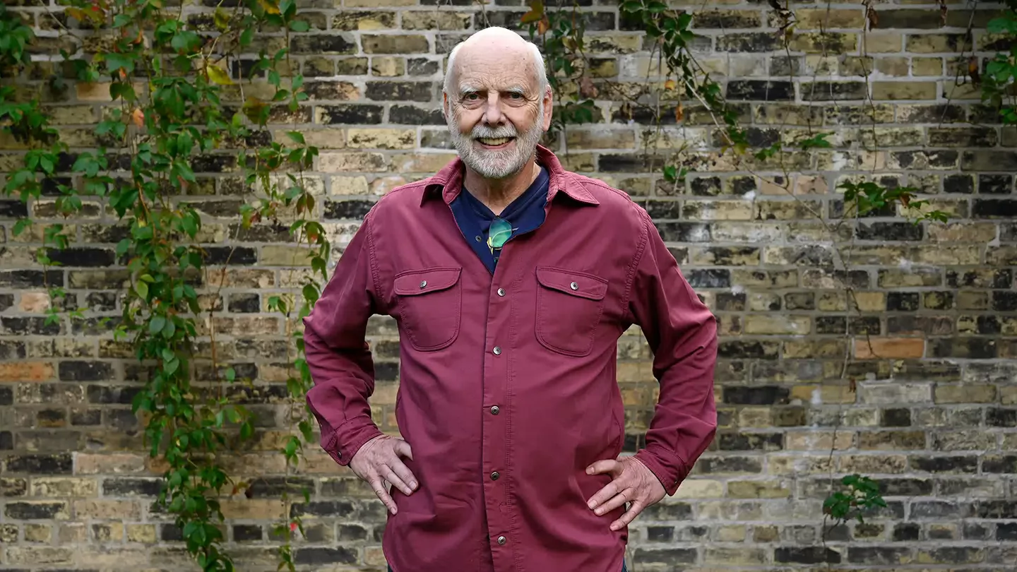 A man with white beard smiling and standing in front of vine covered brick wall
