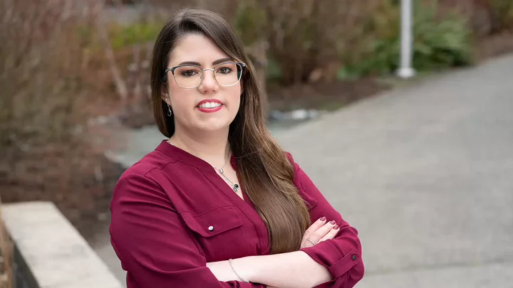 A woman with long hair and glasses standing outside with her arms crossed.