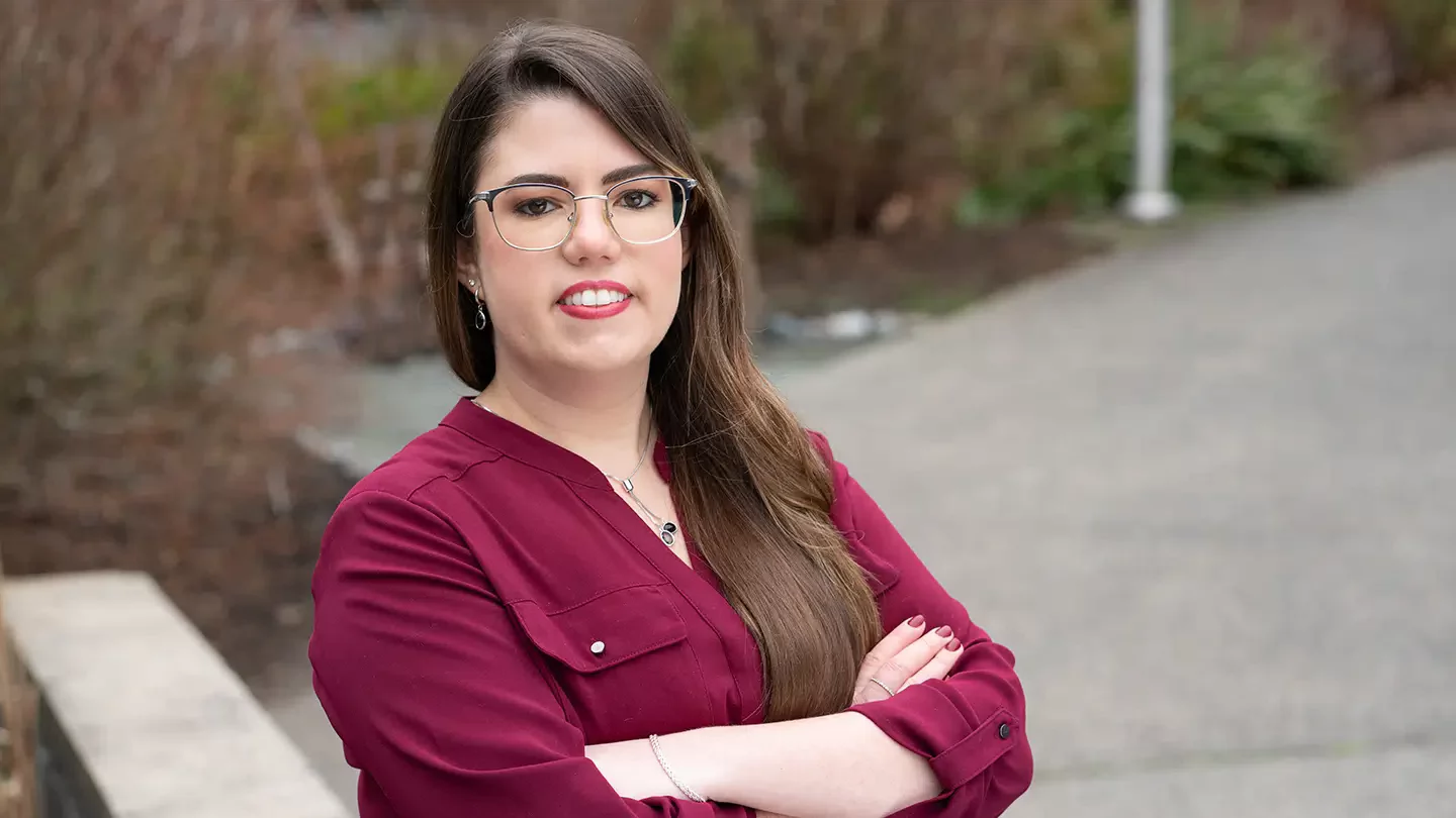 A woman with long hair and glasses standing outside with her arms crossed.