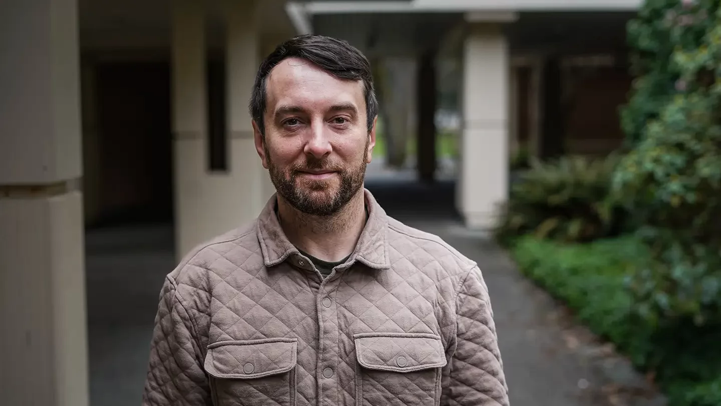 A man with facial hair standing in an outdoor corridor of a building's atrium. 