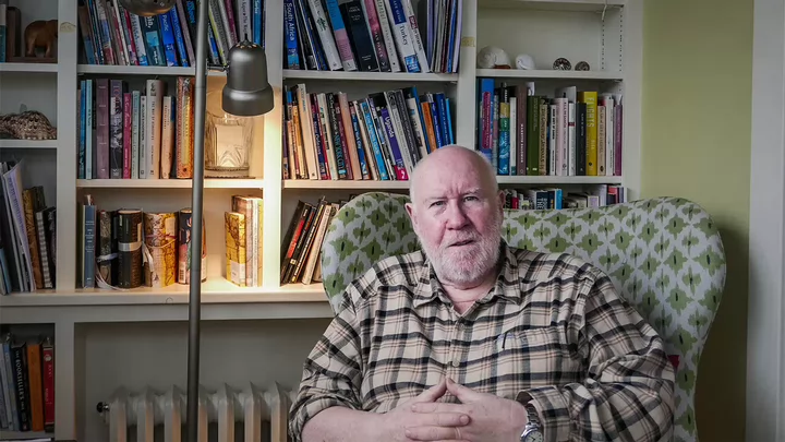 A man in a plaid shirt sitting in an arm chair in front of a book shelf. 
