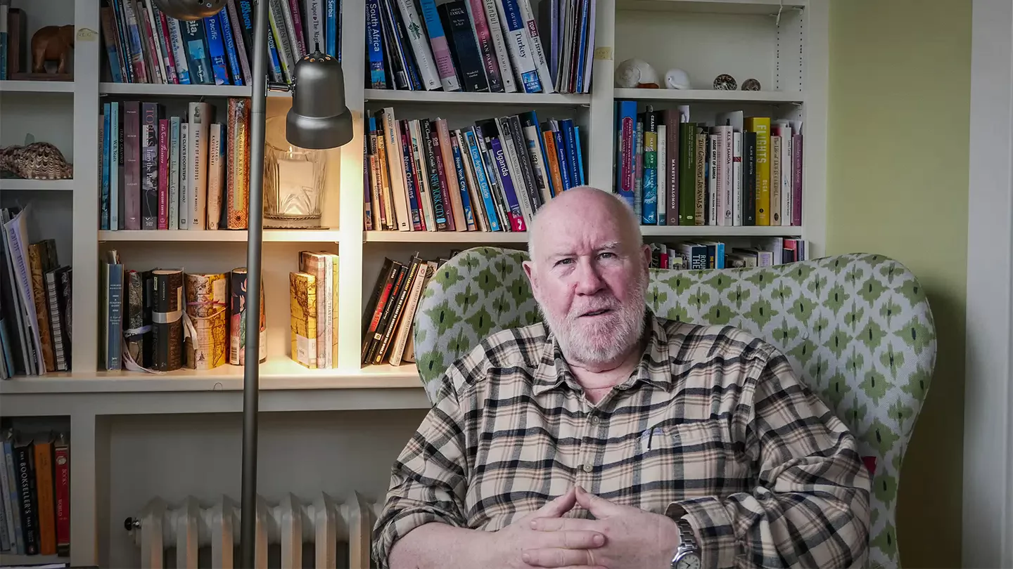 A man in a plaid shirt sitting in an arm chair in front of a book shelf. 