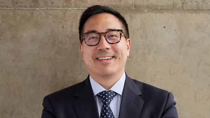 A man smiling and wearing thick rimmed glasses and a jacket and tie. 