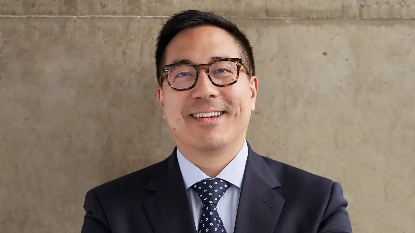 A man smiling and wearing thick rimmed glasses and a jacket and tie. 