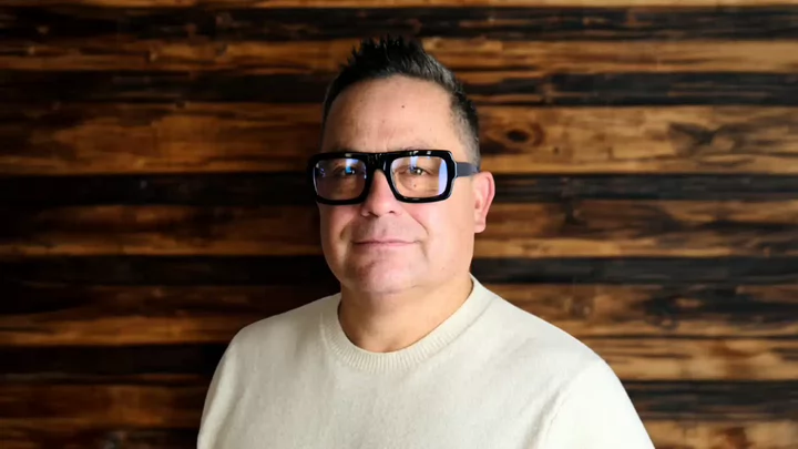 A man with short hair and thick rimmed glasses standing in front of a wooden backdrop.
