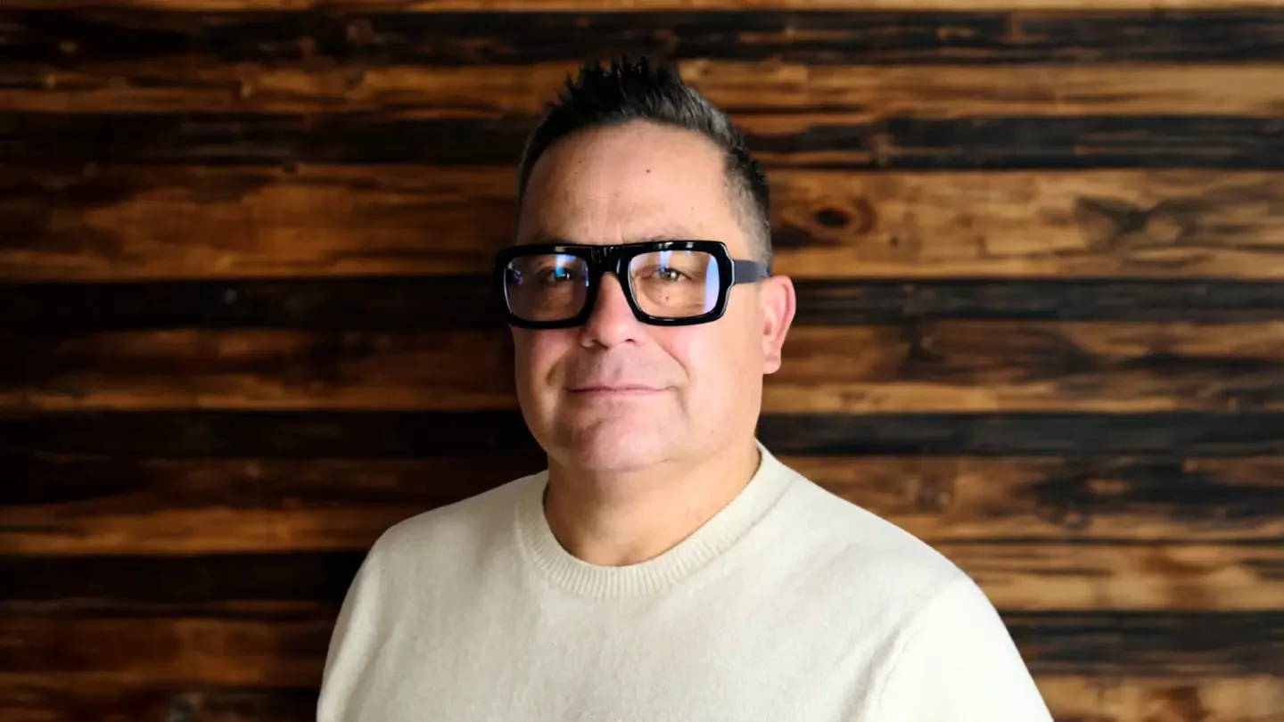 A man with short hair and thick rimmed glasses standing in front of a wooden backdrop.
