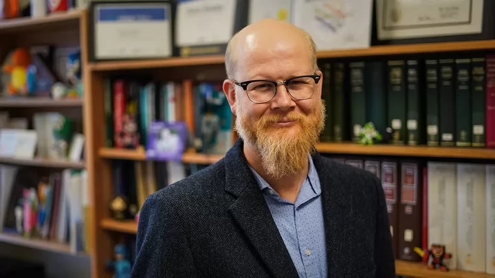 A man with a beard and glasses standing in front of a bookshelf containing books, figurines and framed awards.