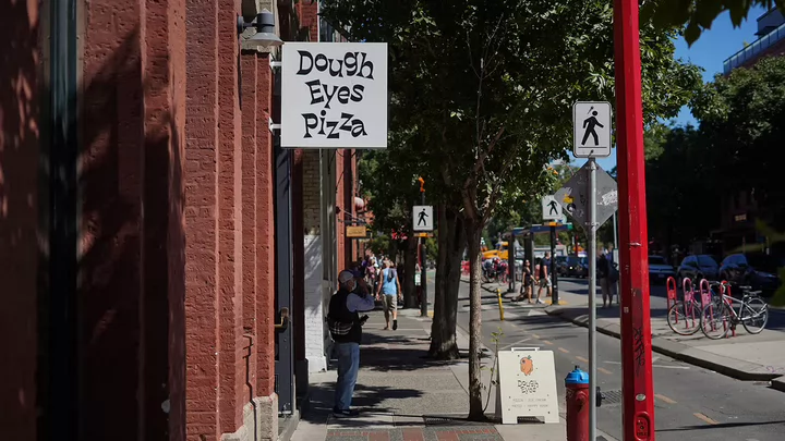 A person stands on the sidewalk under the sign of Dough Eyes restaurant.