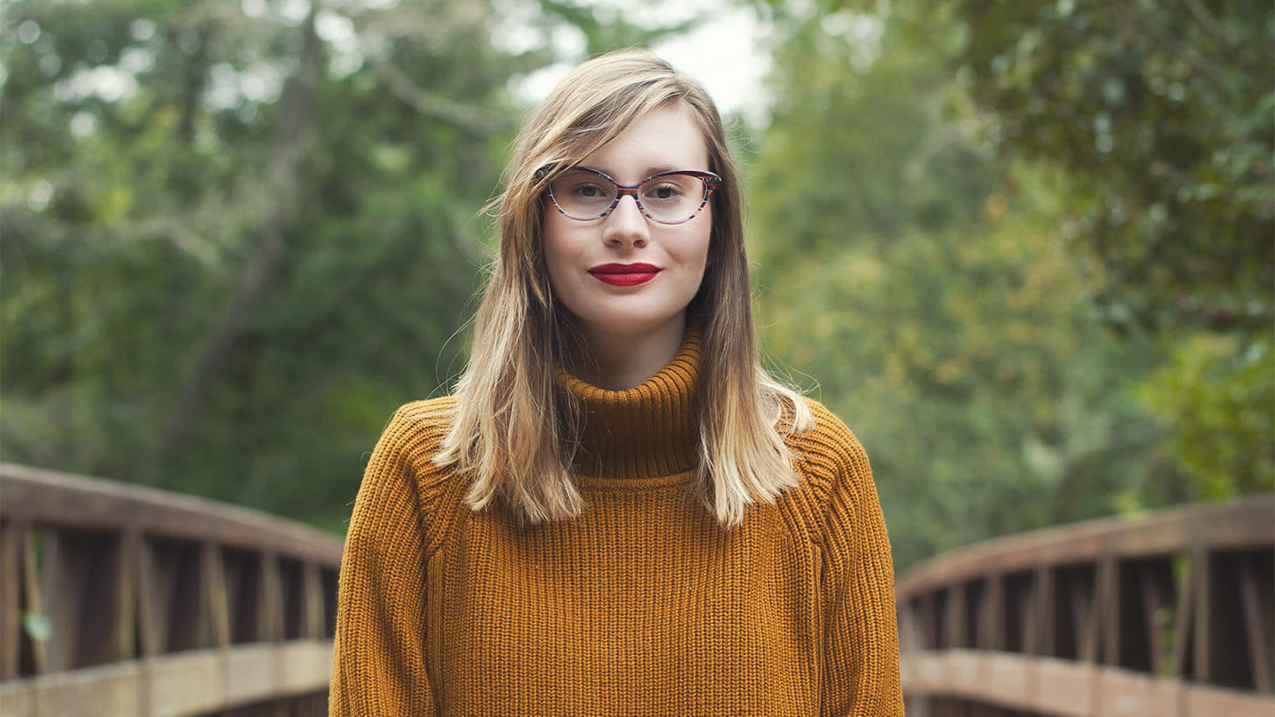 A profile of a woman wearing glasses in an orange sweater on a bridge. 