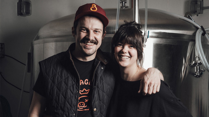 Man with a moustache and baseball hat smiling with his arm around a woman smiling and standing in front of a distilling equipment. 