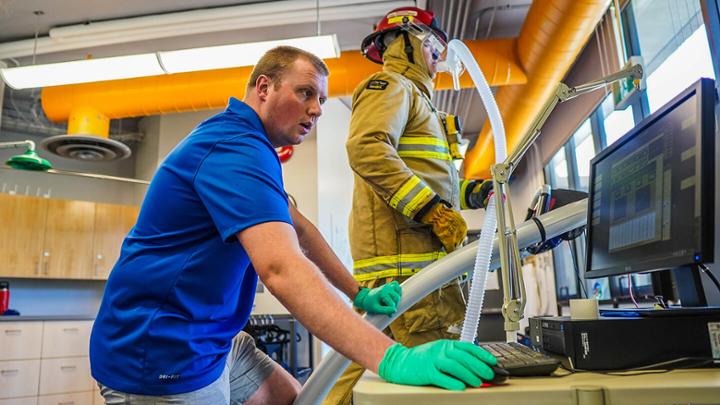 Man looks at computer screen while in the background a firefighter in full gear walks on a treadmill. 