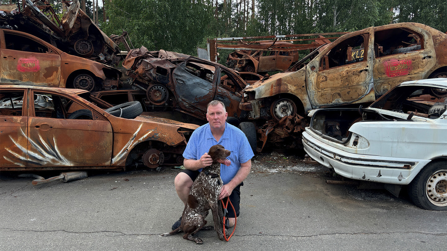 Man kneeling in a car junkyard next to a brown and white dog.