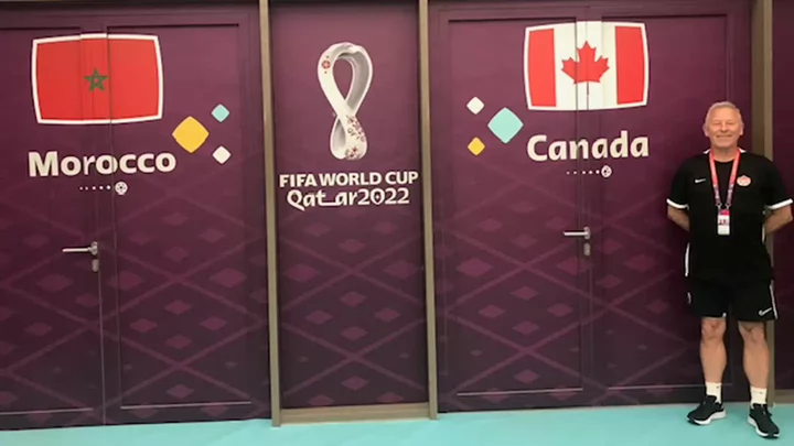 A man wearing athletic shorts, T-shirt and lanyard smiling and standing in front of doors that say Morocco on one side and Canada on the other and Fifa World Cup Qatar 2022 in the middle.  