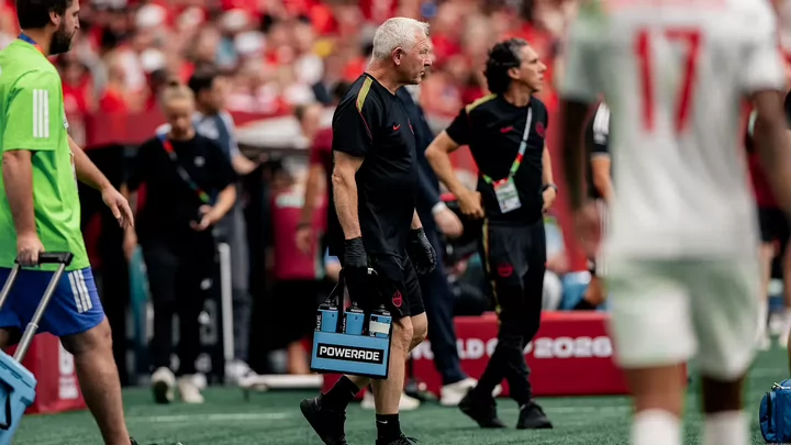 A man with white hair wearing a black T-shirt and shorts walks on a sports field during a game carrying bottles of Powerade..