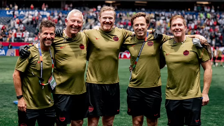 Six people in sports uniforms standing side by side and smiling from the field inside a stadium. 