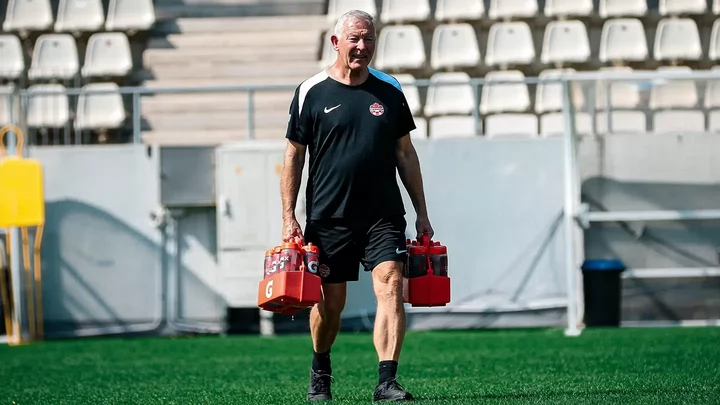 A man with white hair wearing a black T-shirt and shorts walks on a sports field carrying bottles of Gatorade.
