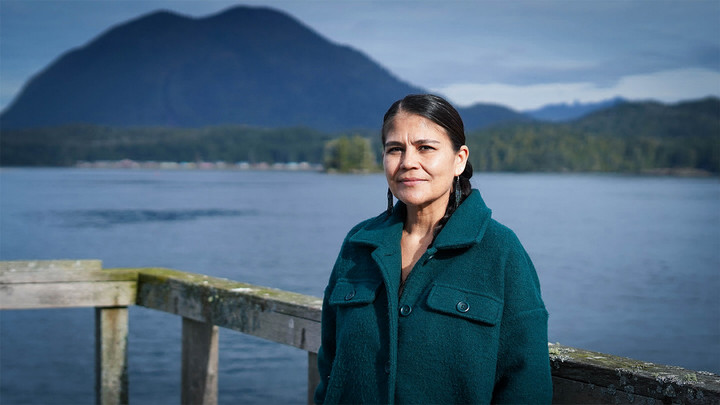 Woman in green jacket standing on a seaside dock with a mountain in the background.