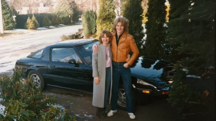 An old photo from the 1970s or 1980s of a man in a leather jacket standing arm in arm with a woman in front of a sports car.